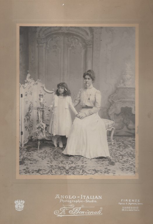 Bella e grande fotografia della fine dell'800, applicata editorialmente su cartoncino intestato al fotografo (Anglo-Italian Photographic Studio F. Benvenuti, Firenze Piazza S. Marco 6). Mm. 285x226 (la foto), mm. 430x335 (il cartone). Raffigura una giovane madre seduta con la piccola figlia accanto, in un ambiente neorococò. Alcune consunzioni al volto della madre, lievi mancanze al margine bianco della foto.